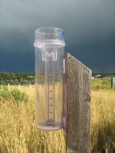 A photograph of a CoCoRaHS rain gauge. The gauge is attached to a 4-6 foot wood pole located in an open field. Dark storm clouds are brewing in the background.